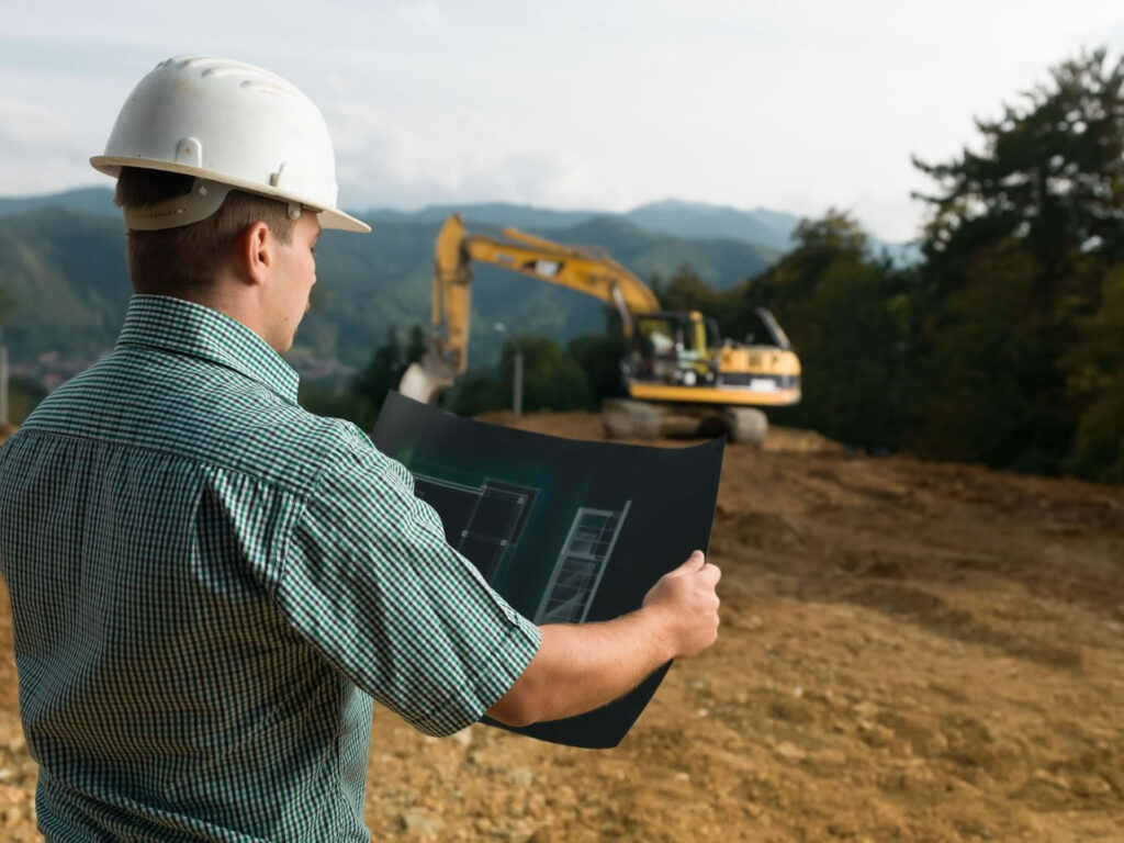 Construction worker reviewing grading plans on a job site with excavator in the background.